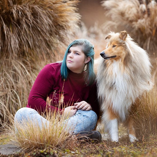 Eine junge Frau, mit kurzen blauen Haaren und einem roten Oberteil sitzt neben einem sable Langhaar Collie. Beide schauen in die Ferne
